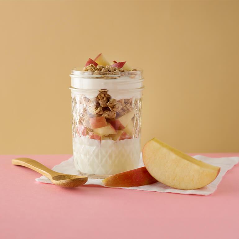 A layered parfait in a glass jar with yogurt, diced apples, and granola. It sits on a napkin with a wooden spoon beside it on a pink surface. A slice of apple is placed next to the jar. The background is a warm, light brown color.