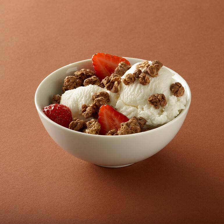 A white bowl filled with scoops of vanilla ice cream, topped with clusters of granola and slices of fresh strawberries, sits on a brown surface.